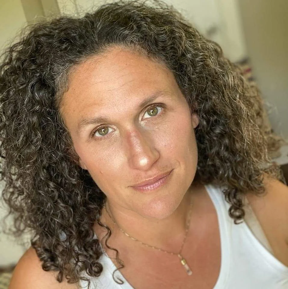 Close-up of a woman with curly brown hair, wearing a white tank top, gold necklace, and smiling softly at the camera.