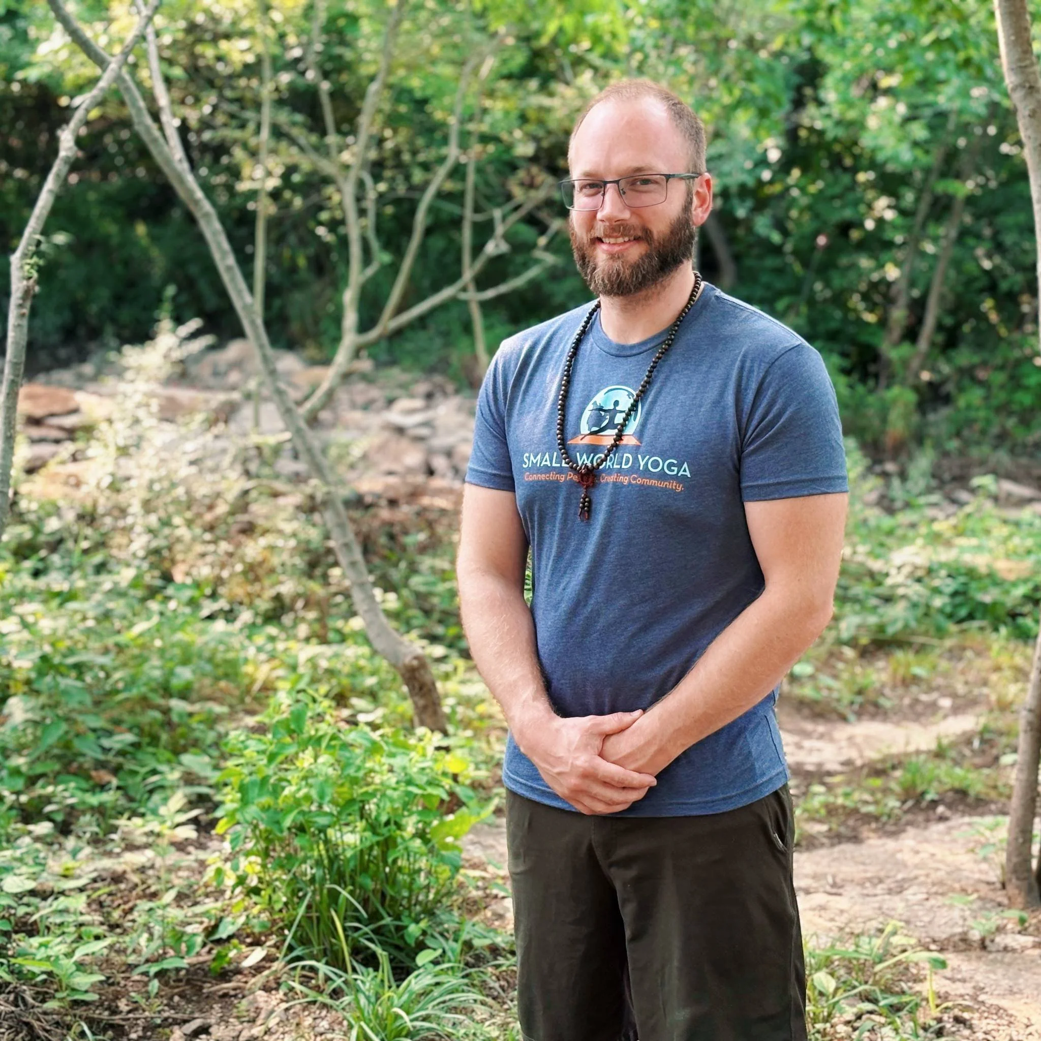 Man with glasses and a beard, wearing a blue Small World Yoga T-shirt, standing outdoors in a wooded area with green foliage.