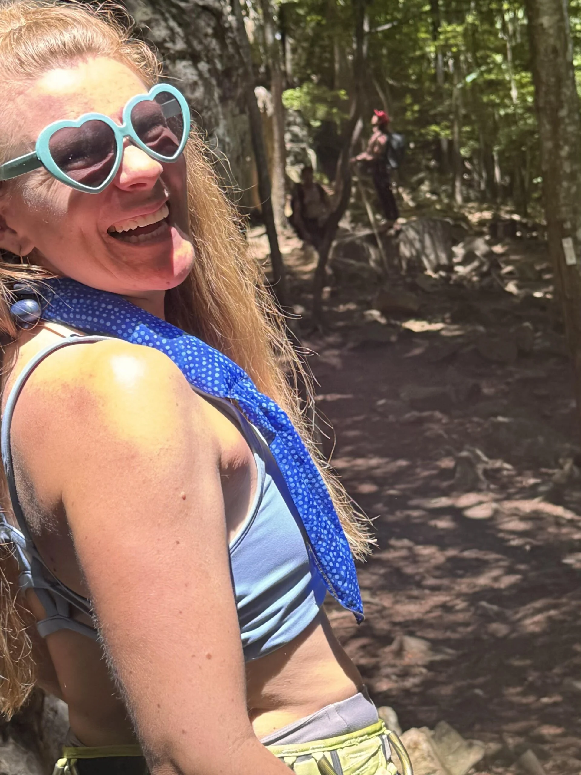 A woman with red hair, wearing heart-shaped sunglasses, smiling, outdoors in a wooded area on a sunny day.