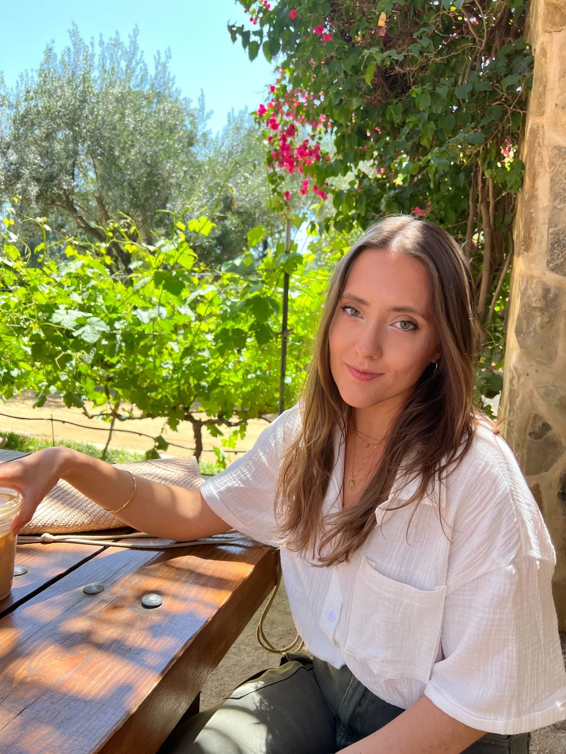 A woman with long brown hair and a white shirt sitting at a wooden table outdoors, with greenery and pink flowers in the background.