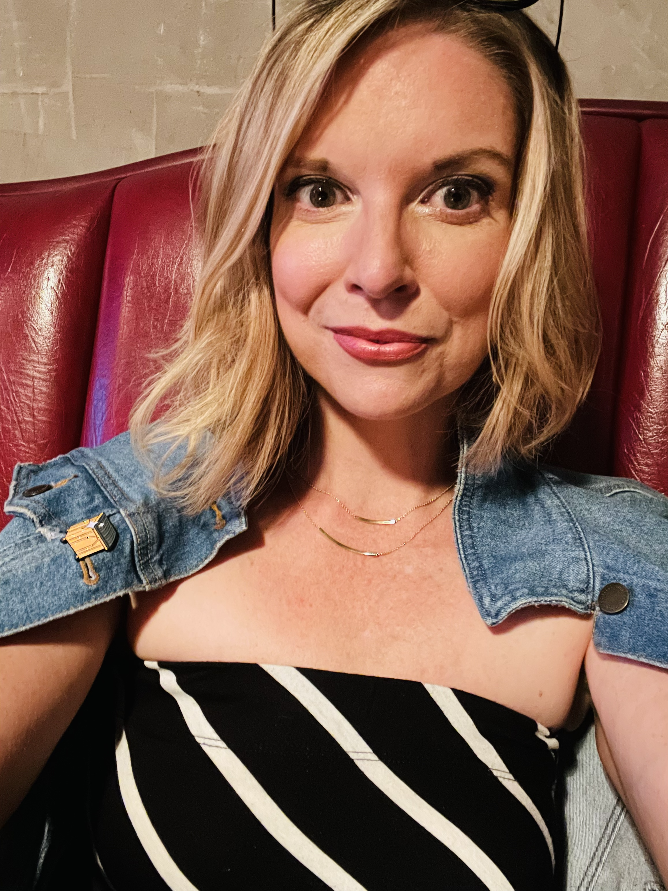 A woman with shoulder-length blonde hair, wearing a striped black and white top and a small denim jacket with a shoulder pin, sits on a red leather seat, smiling at the camera.