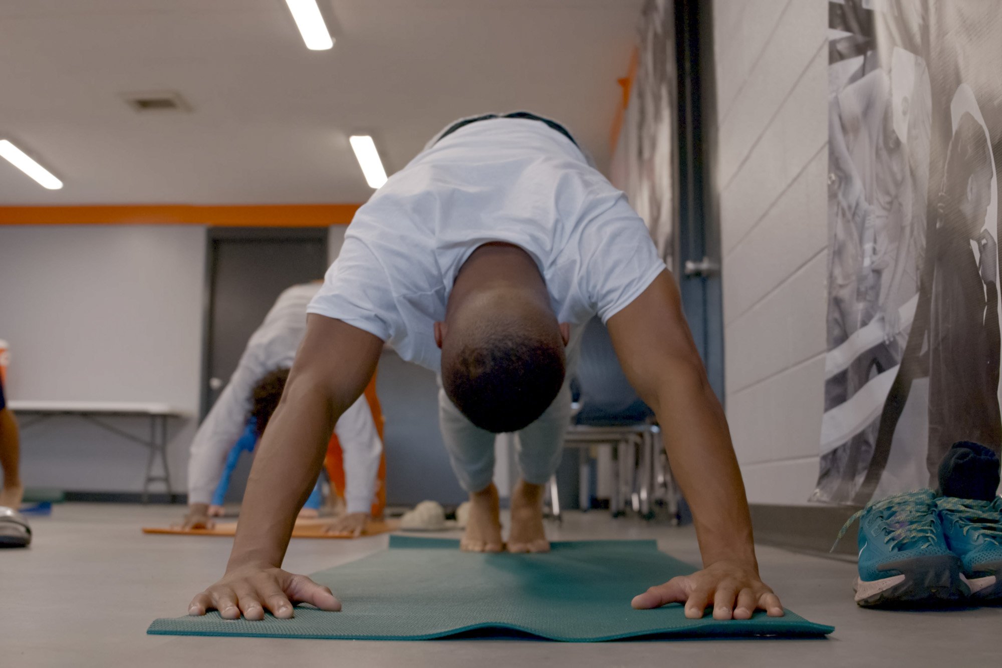 Man in white T-shirt practicing yoga in a plank position on a green yoga mat in a room with other people.