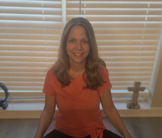 A woman with long, wavy hair smiling while sitting in front of window blinds, wearing a red T-shirt.