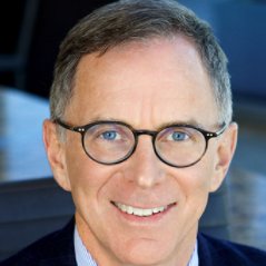 Close-up of a smiling man with glasses, short brown hair, wearing a suit and tie.