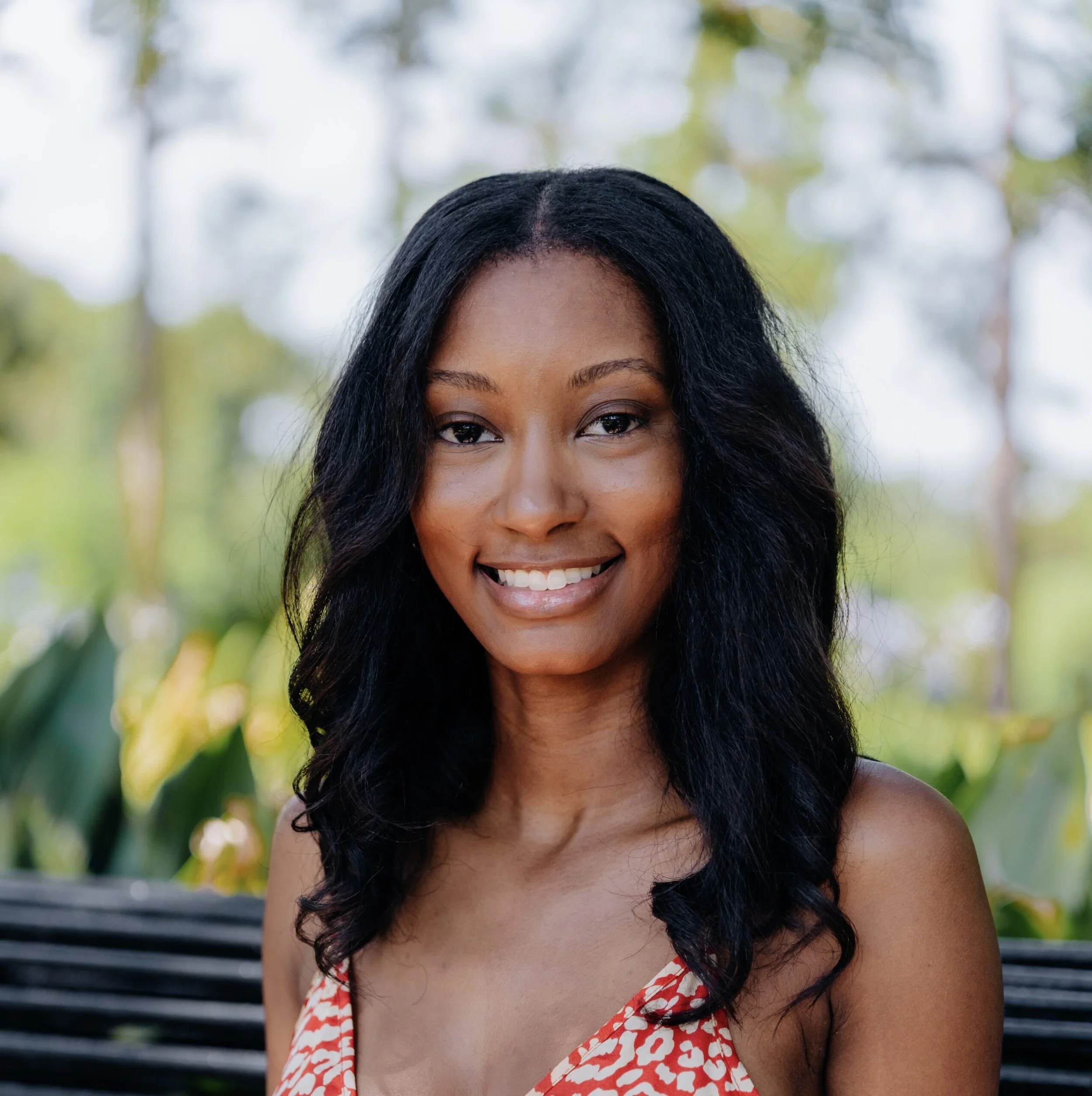 A young woman with long, dark wavy hair smiling outdoors with green trees and blurred background.