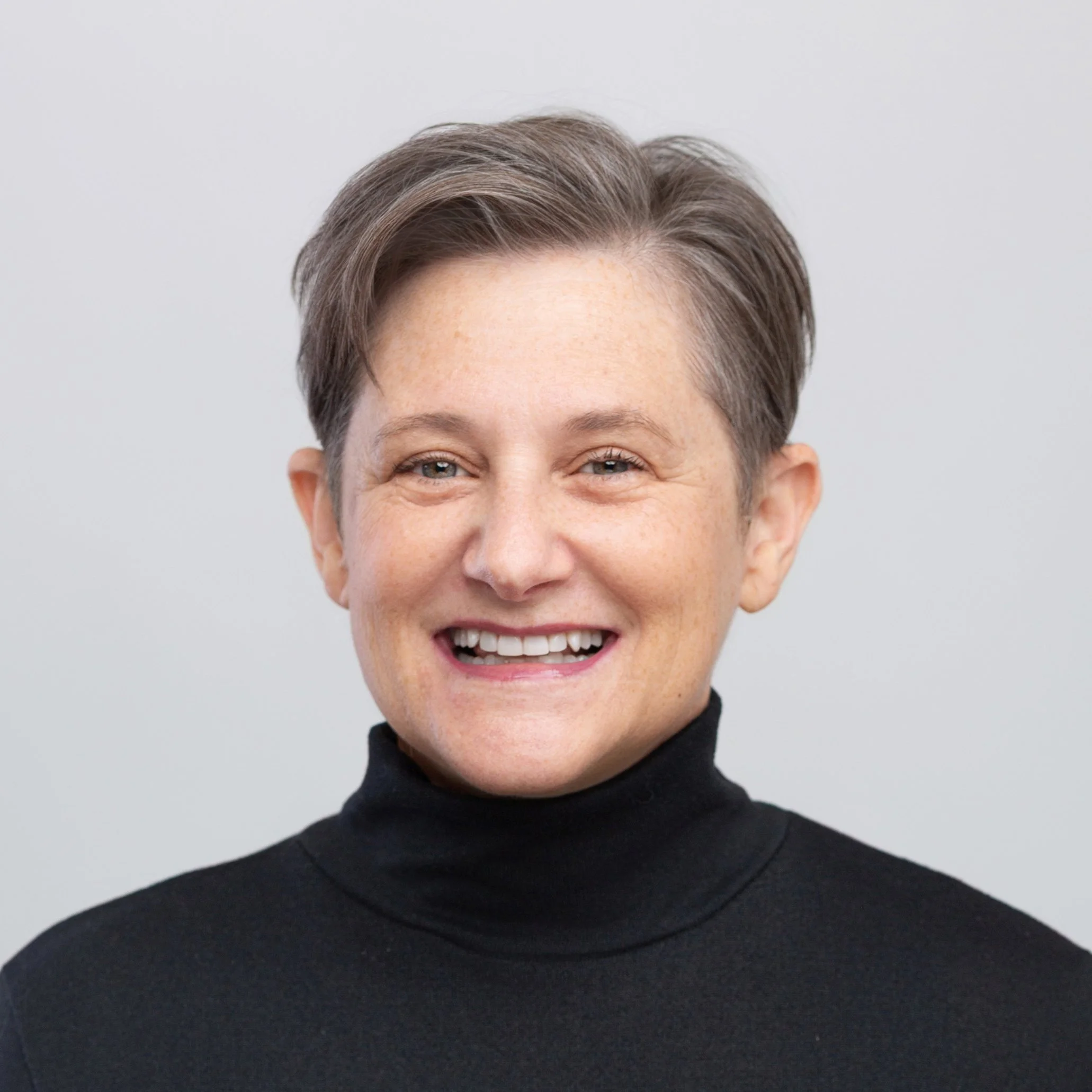 Close-up of a woman with short, gray hair, smiling, dressed in a black turtleneck, against a plain background.