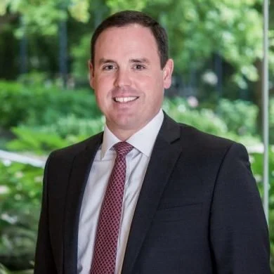 A man in a suit and tie smiling outdoors with a blurred green park background.