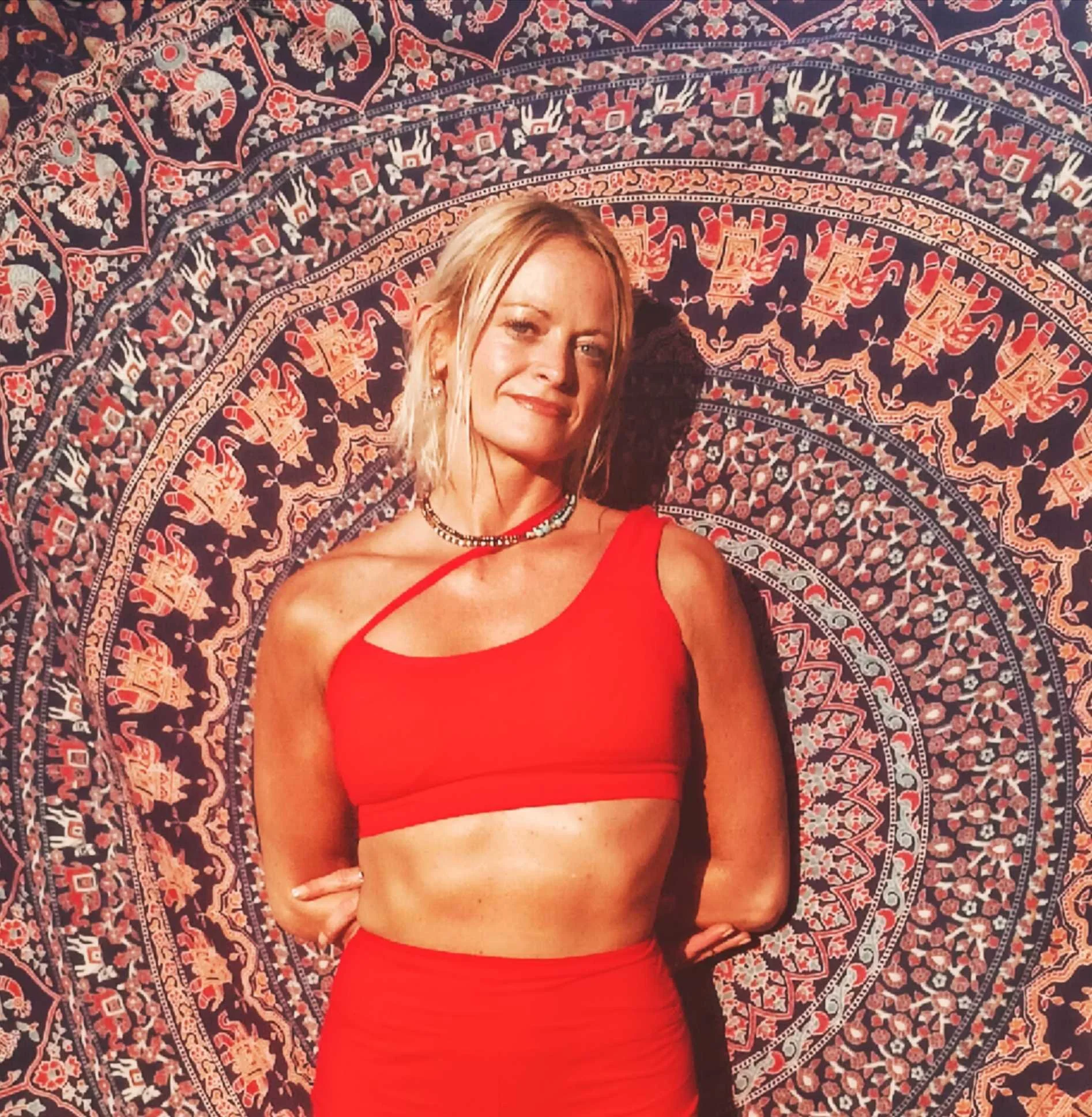 A woman in a red crop top and matching skirt posing in front of a decorative, patterned tapestry with intricate designs.