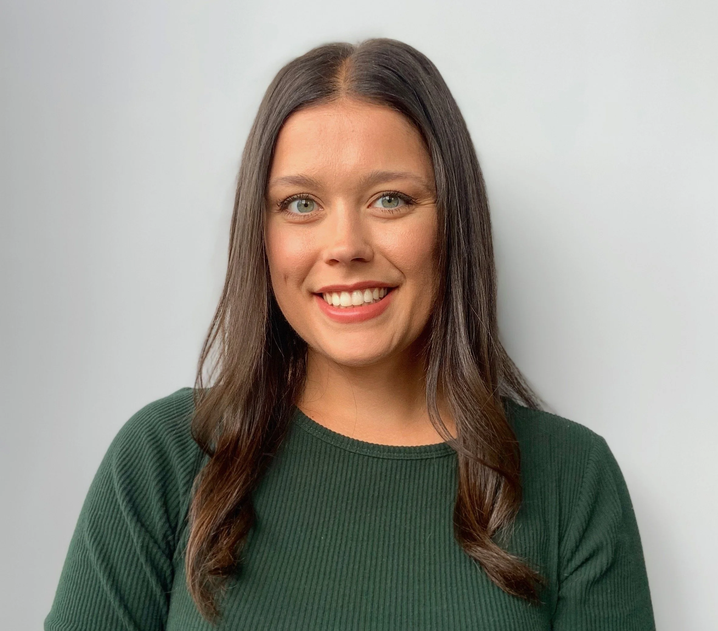 A woman with long dark hair wearing a green top, smiling in front of a plain light gray background.