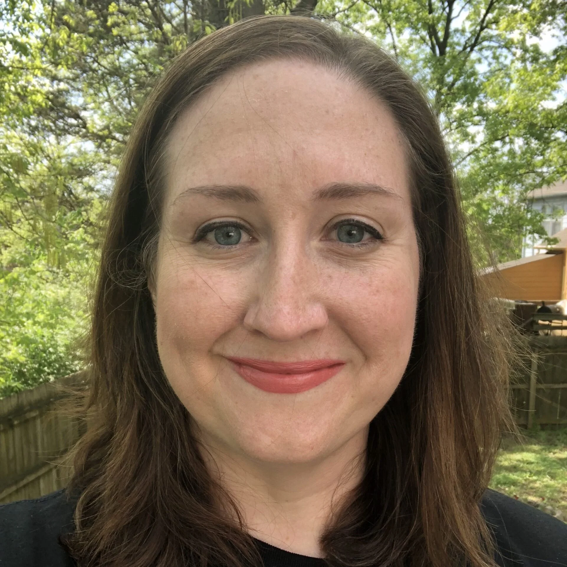 Close-up of a woman with brown hair and fair skin, outdoors with trees and a wooden fence in the background.