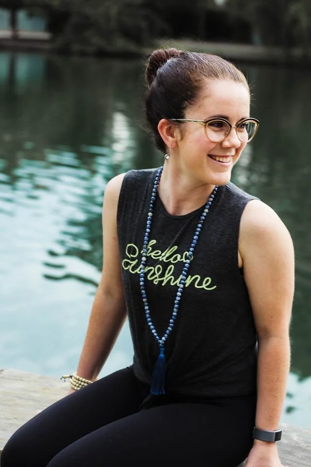 A woman sitting outdoors by a body of water, smiling, wearing glasses, a sleeveless black shirt with green writing, a long blue beaded necklace, and a watch.