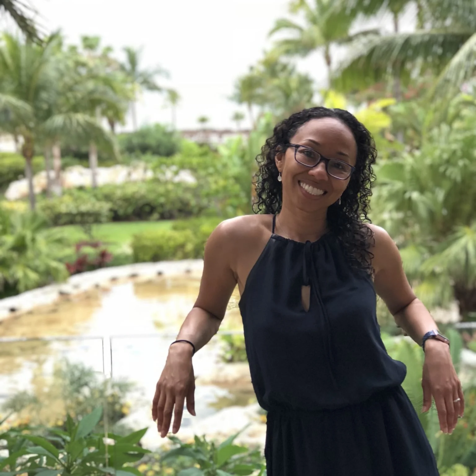 Woman smiling outdoors in a tropical garden with lush green plants and a small pond.