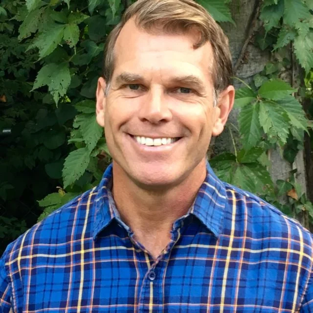 A man smiling outdoors with green leafy background, wearing a blue plaid shirt.