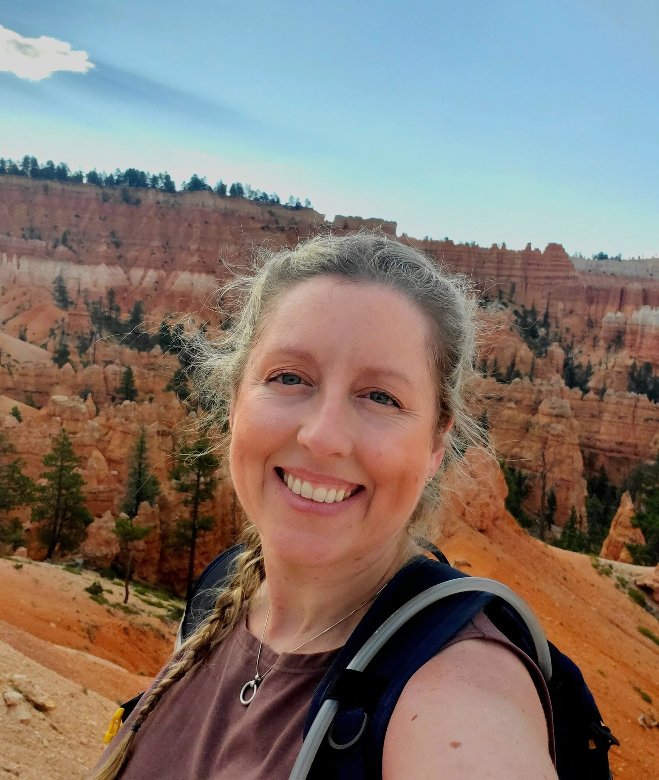 A woman smiling with a braid, wearing a backpack, in front of red rock formations and trees at a scenic outdoor location.
