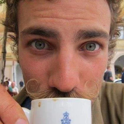 Close-up of a man with curly hair and a mustache drinking from a white mug outdoors.