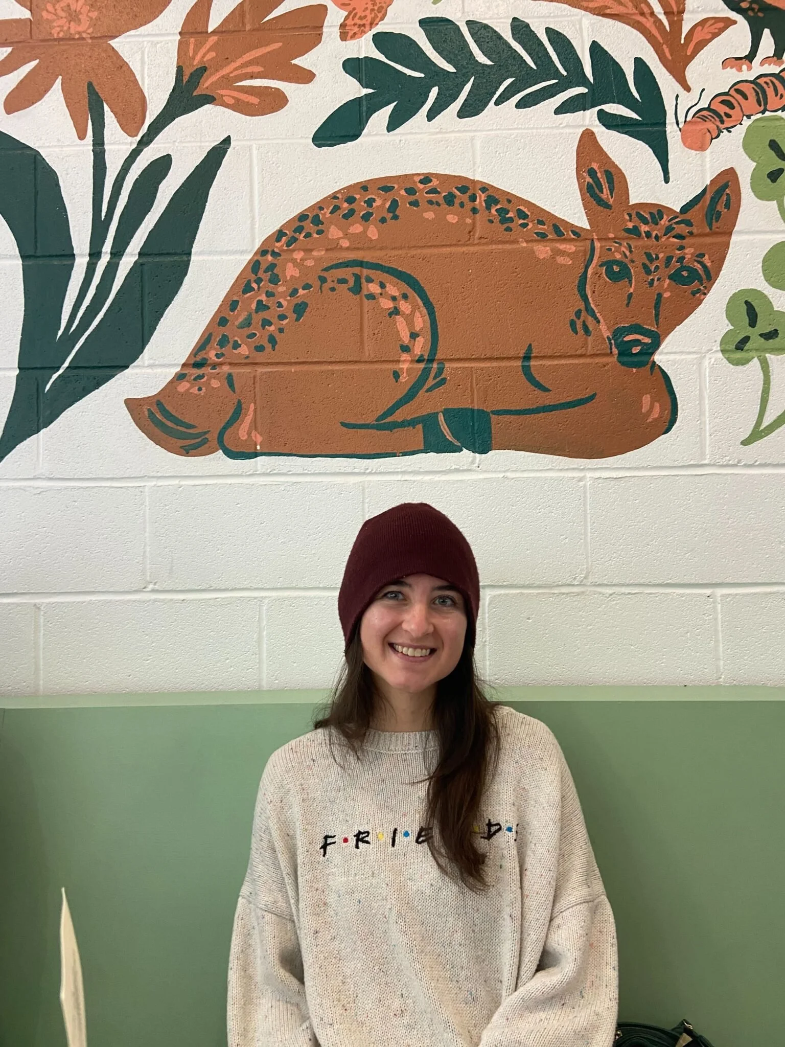A woman with long brown hair wearing a maroon beanie and a beige sweater with the word 'Friends' on it, smiling and sitting in front of a wall with a large painted artwork of a sleeping cheetah surrounded by green and orange plants.