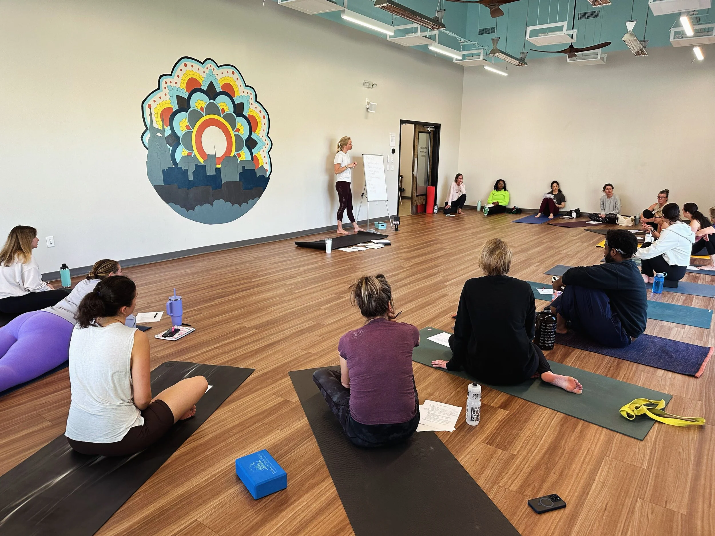 A group of people attending a yoga or meditation class in a spacious, well-lit studio. The instructor is standing on a platform at the front, speaking to the group. Participants are seated on yoga mats, some taking notes or listening attentively.