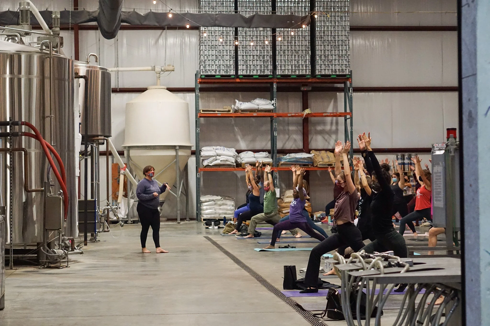 People participating in a yoga class inside a brewery or industrial space, with large brewing tanks on one side and high shelves with stored supplies at the back, practicing poses with arms raised.
