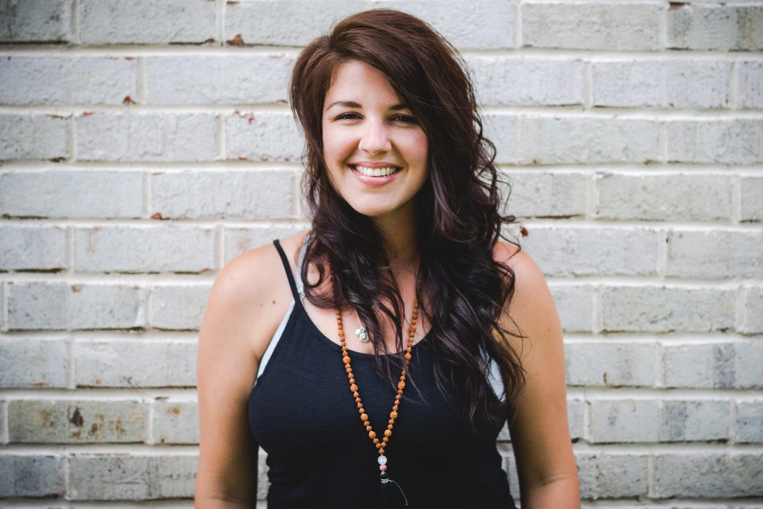 A smiling woman with long, dark, wavy hair stands in front of a white brick wall.