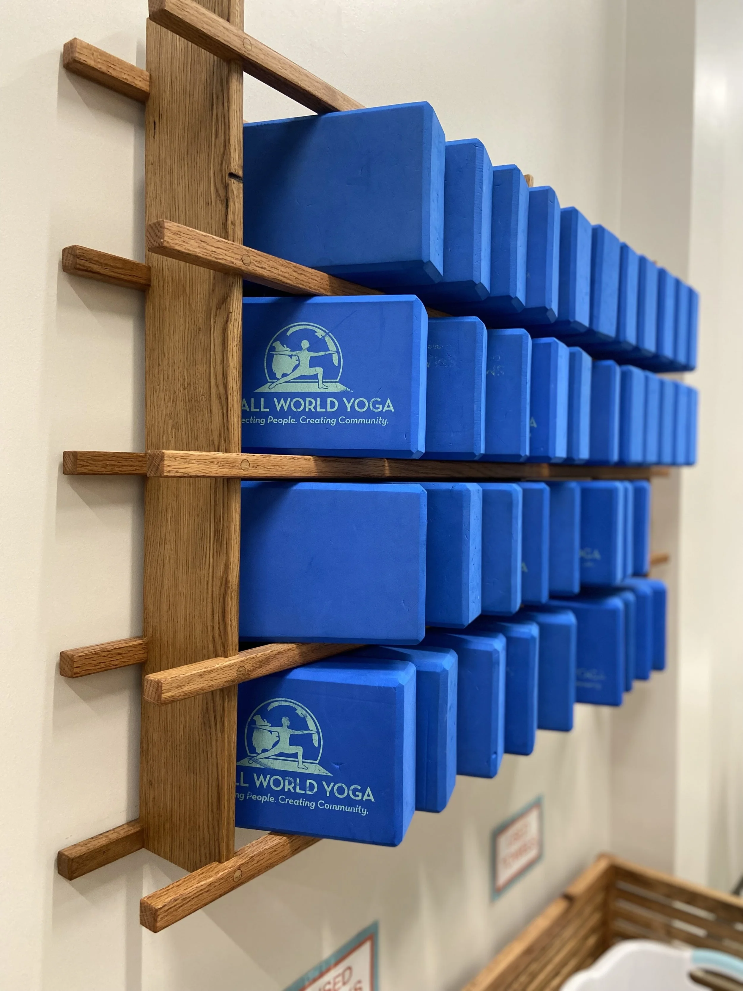 Wall mounted wooden holder with multiple blue yoga blocks. The blocks have a logo and the words "ALL WORLD YOGA" on them.