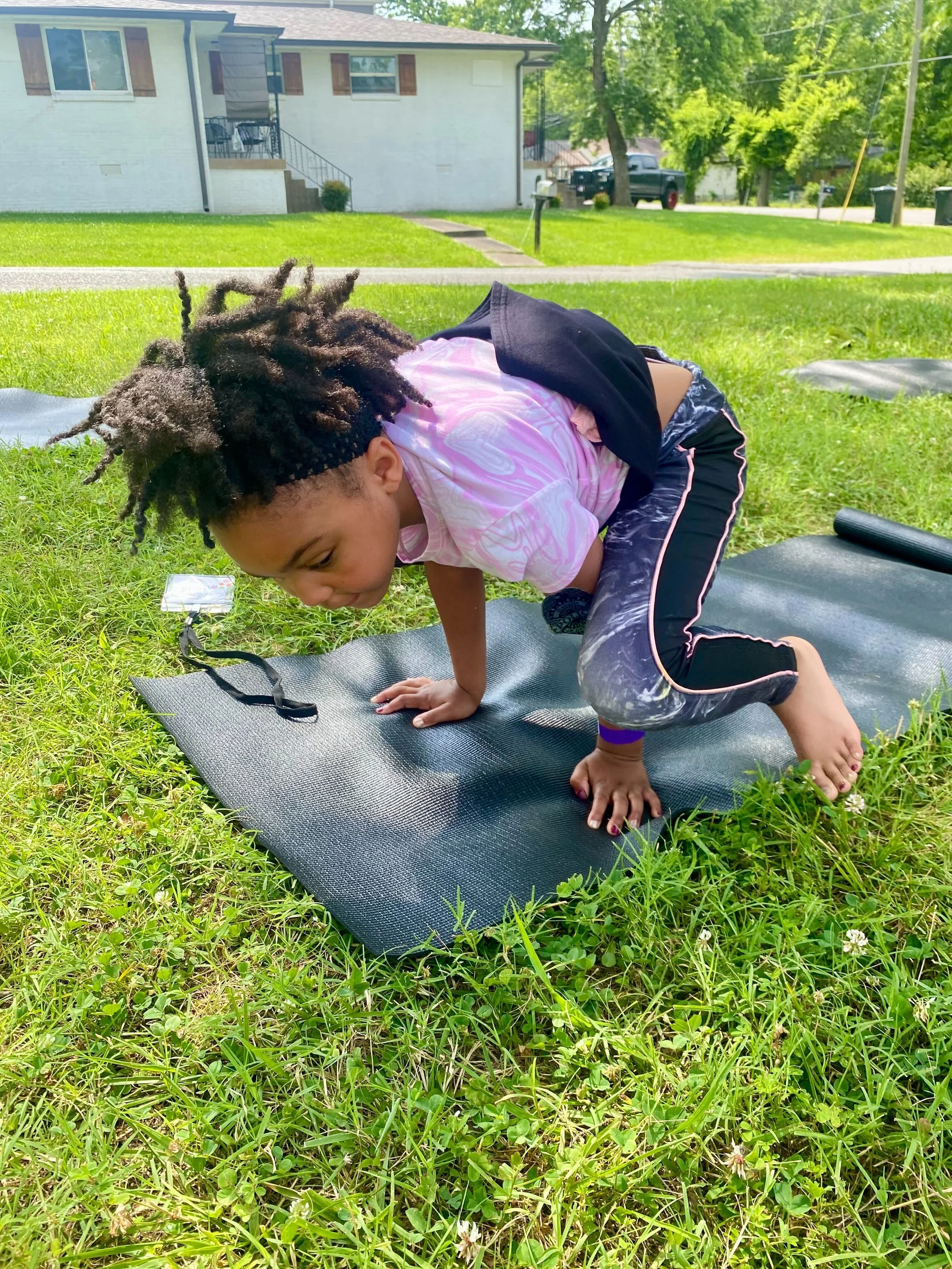 A young girl with curly hair doing a yoga pose on a black mat outdoors on a grassy lawn, with houses and trees in the background on a sunny day.