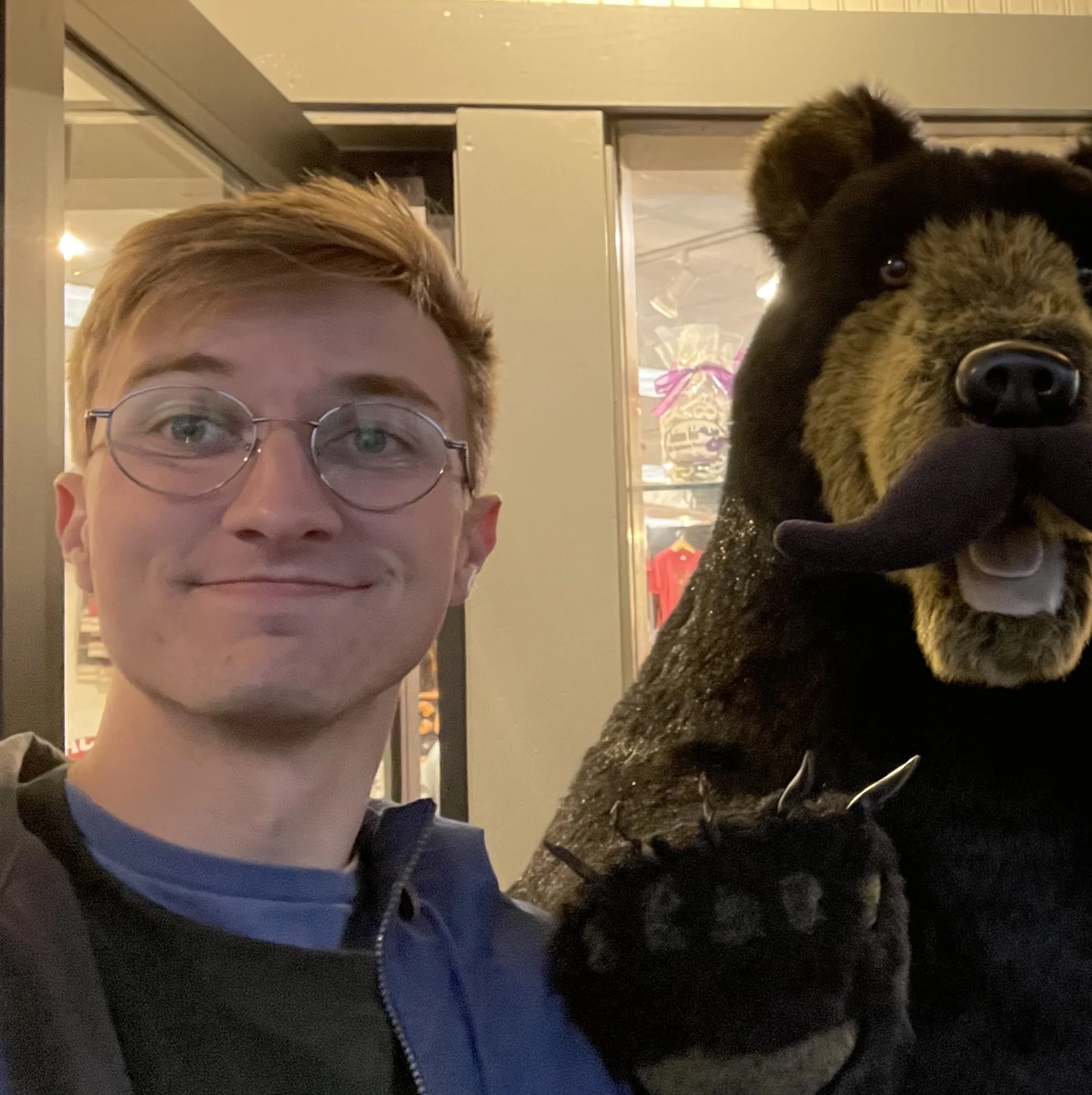 A young man with glasses taking a selfie next to a large, black and brown bear costume with a tongue sticking out.