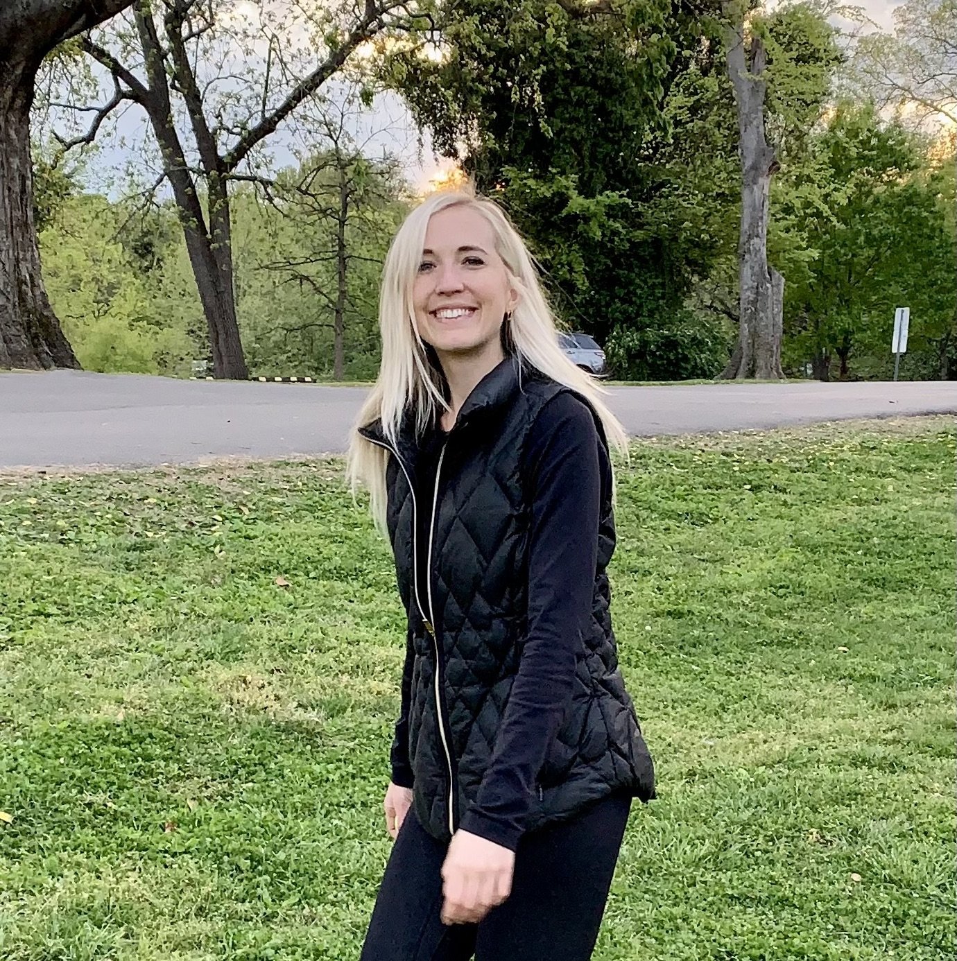 A woman with blonde hair wearing a black quilted vest and black pants, smiling outdoors on a grassy area with trees and a road in the background during sunset.