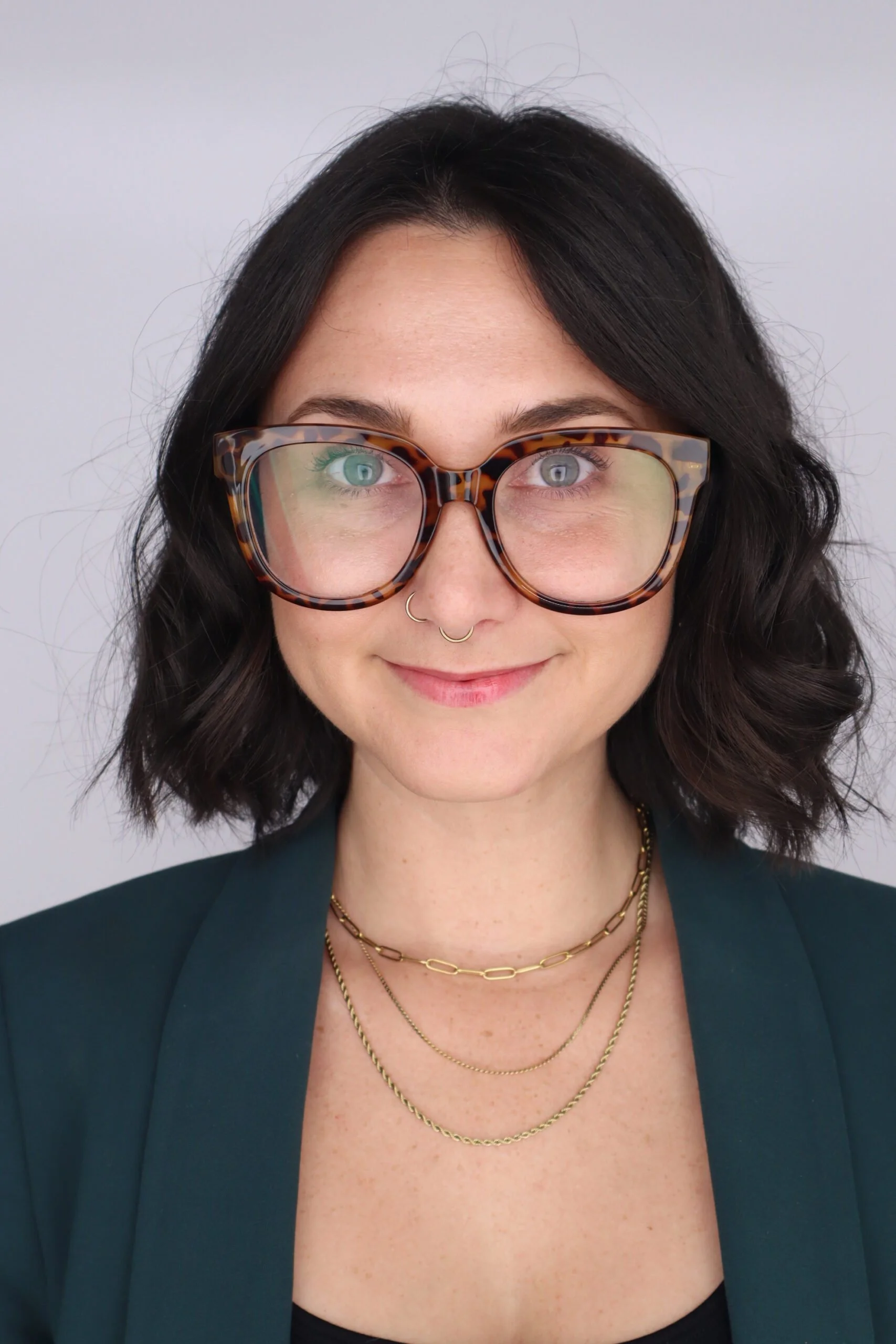 A woman with dark wavy hair, wearing large tortoiseshell glasses, a nose ring, multiple gold necklaces, and a dark green blazer, smiling at the camera.