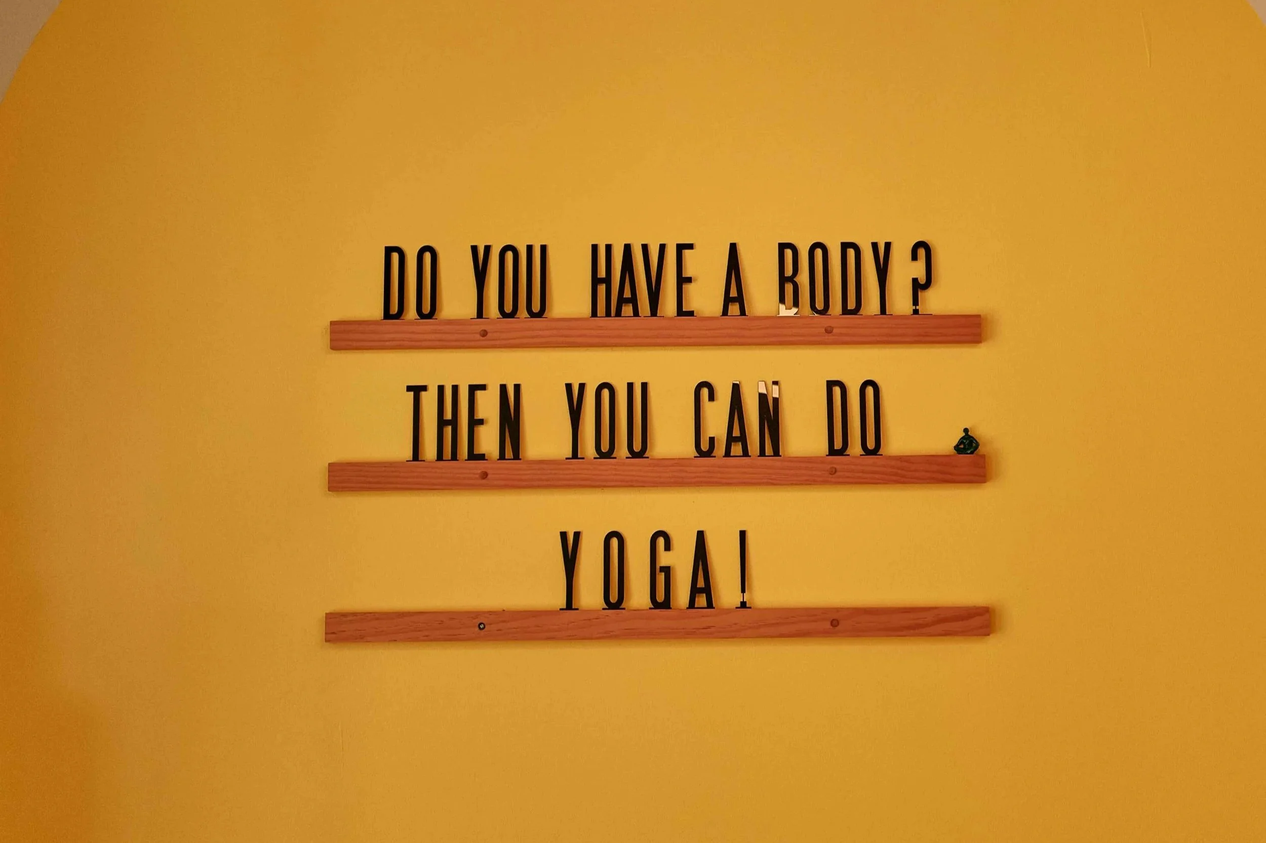 A yellow wall with black lettering spelling out a quote about bodies and yoga, arranged with wooden lines separating parts of the text.