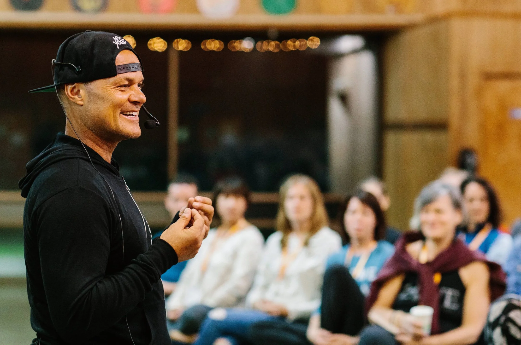 A man wearing a black hoodie and a backwards cap with a microphone headset, smiling and speaking to an audience in a room with wooden walls and blurred people seated in the background.