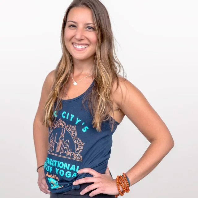 Smiling woman with long blonde hair wearing a navy blue tank top with yoga-themed text and designs, standing against a plain light background.