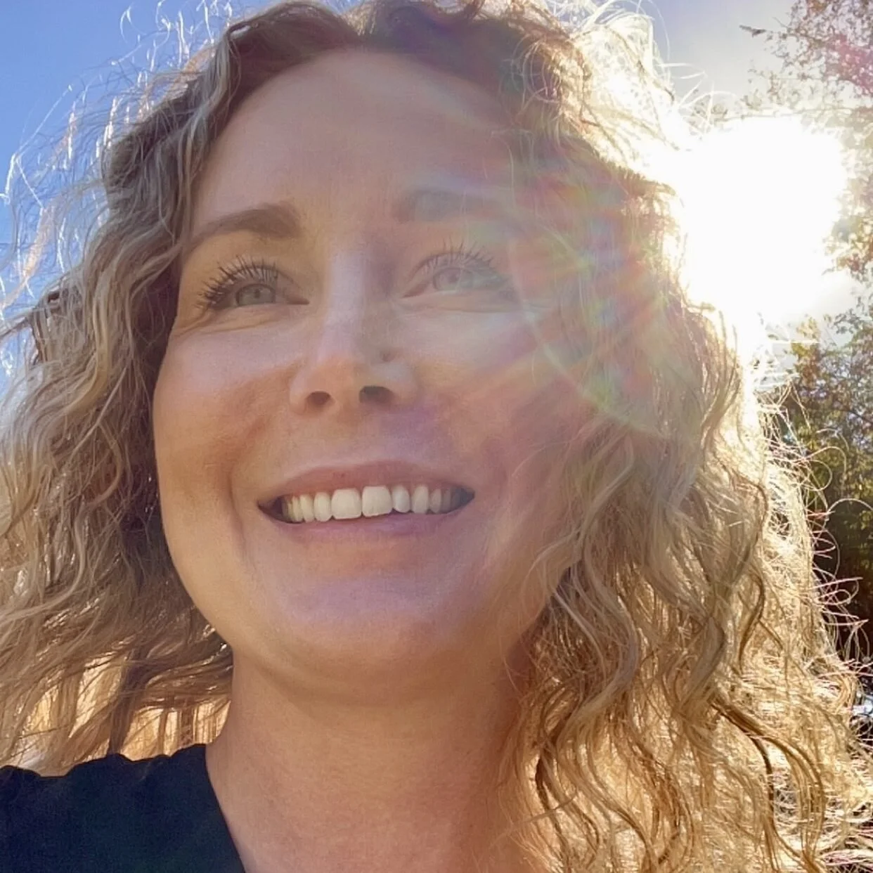 A woman with curly blonde hair smiling outdoors in bright sunlight, with lens flare and a blue sky background.