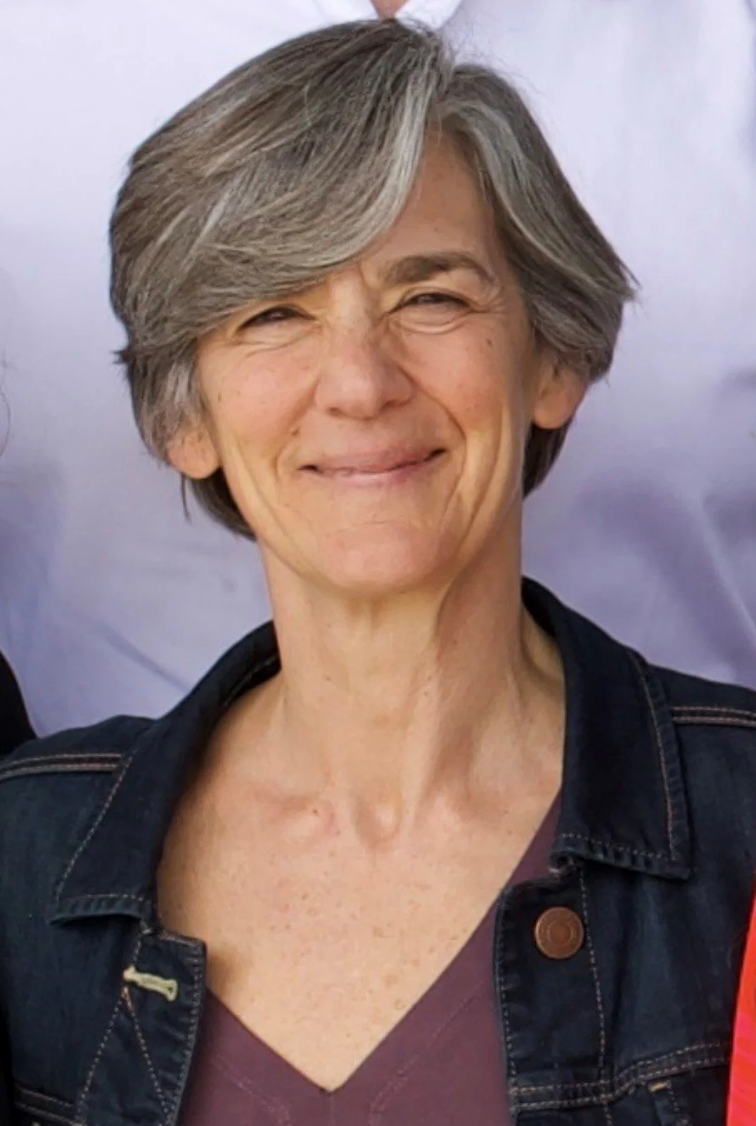 A smiling woman with short, gray hair wearing a dark denim jacket and a maroon top.