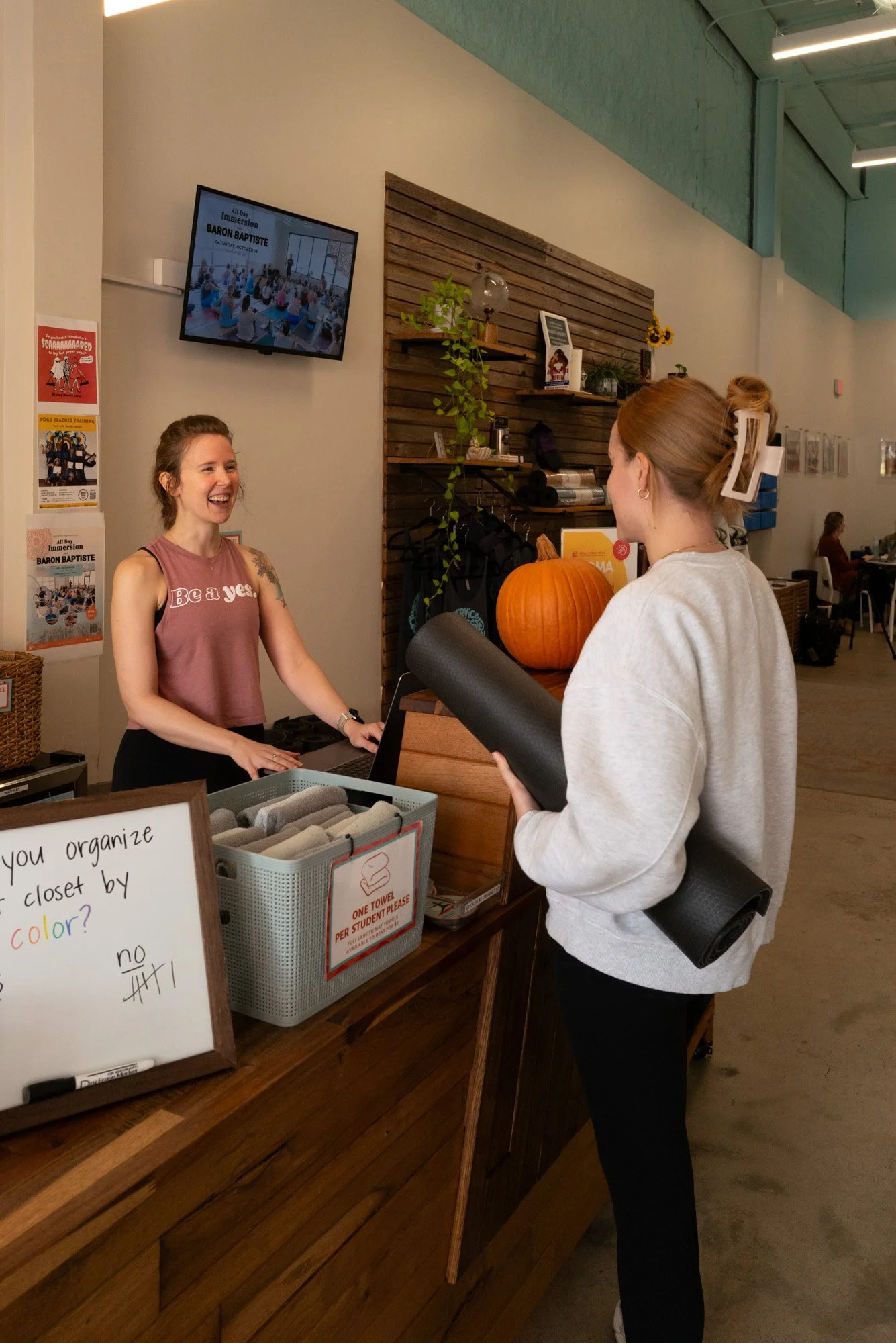 A woman with blonde hair tied back, holding a black yoga mat, stands at a reception counter, smiling and speaking to a receptionist with long dark hair, wearing a sleeveless pink top. The reception area has a pumpkin on the counter and a sign that re