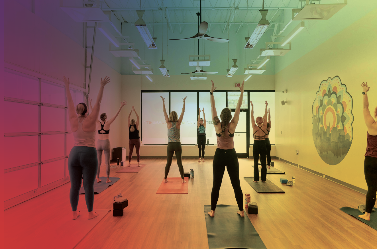 A group of people participating in a yoga class in a spacious, well-lit room with wooden flooring, colorful walls, and large windows at the front.