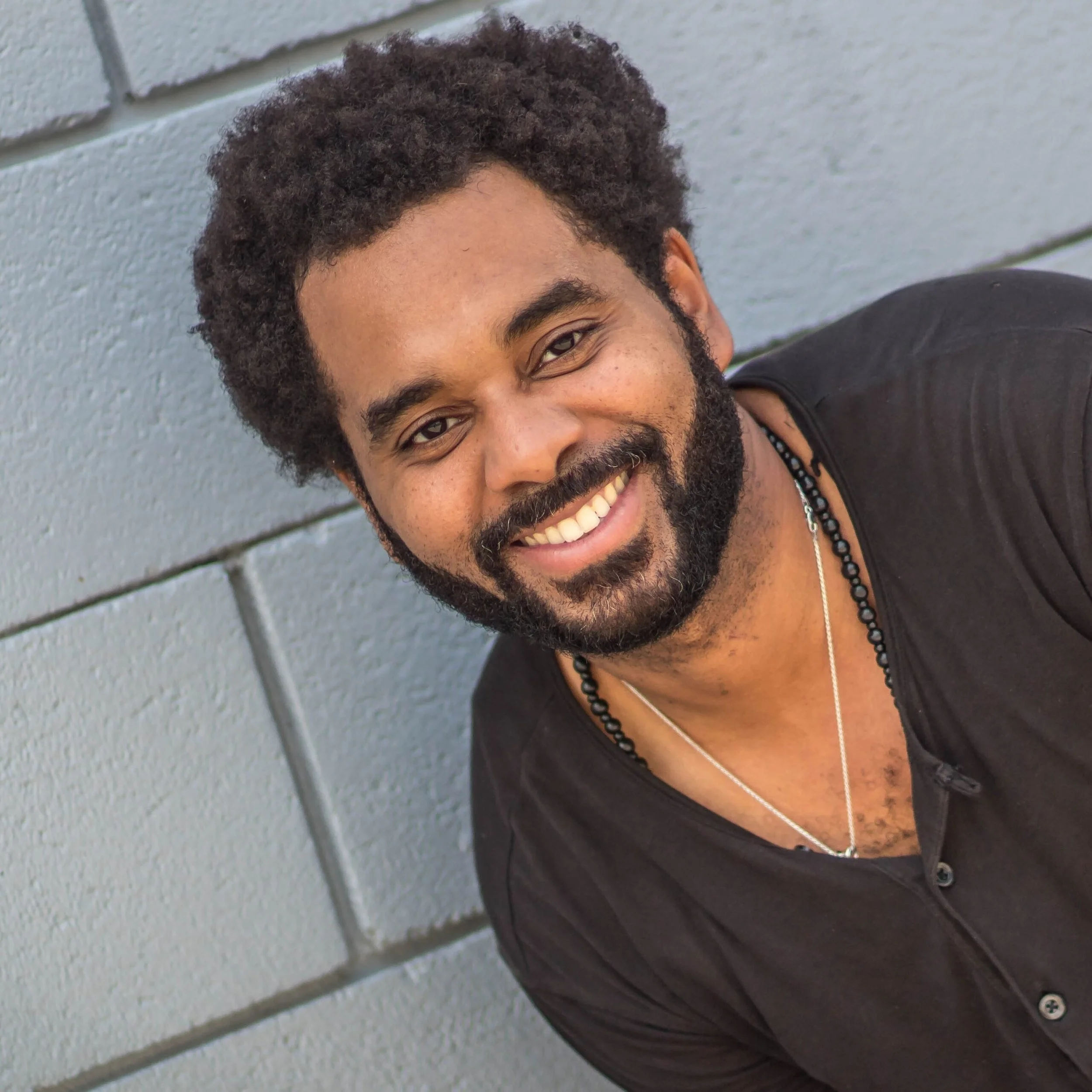 A smiling man with curly hair and a beard, wearing a black shirt and layered necklaces, leaning against a grey brick wall.