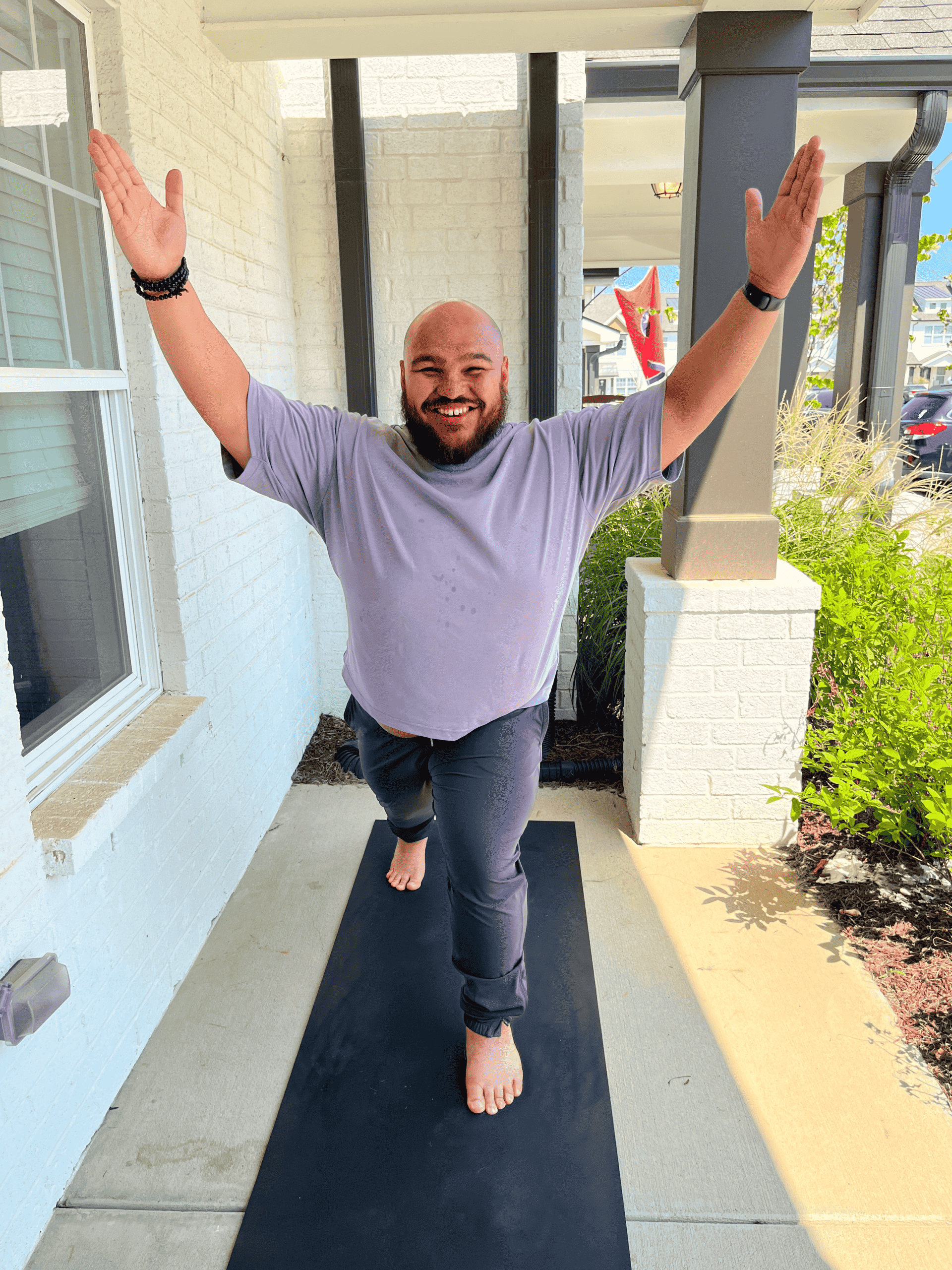 A man practicing yoga on a black mat outside on a porch, standing on one leg with arms raised, smiling, wearing a grey t-shirt, dark pants, and no shoes.