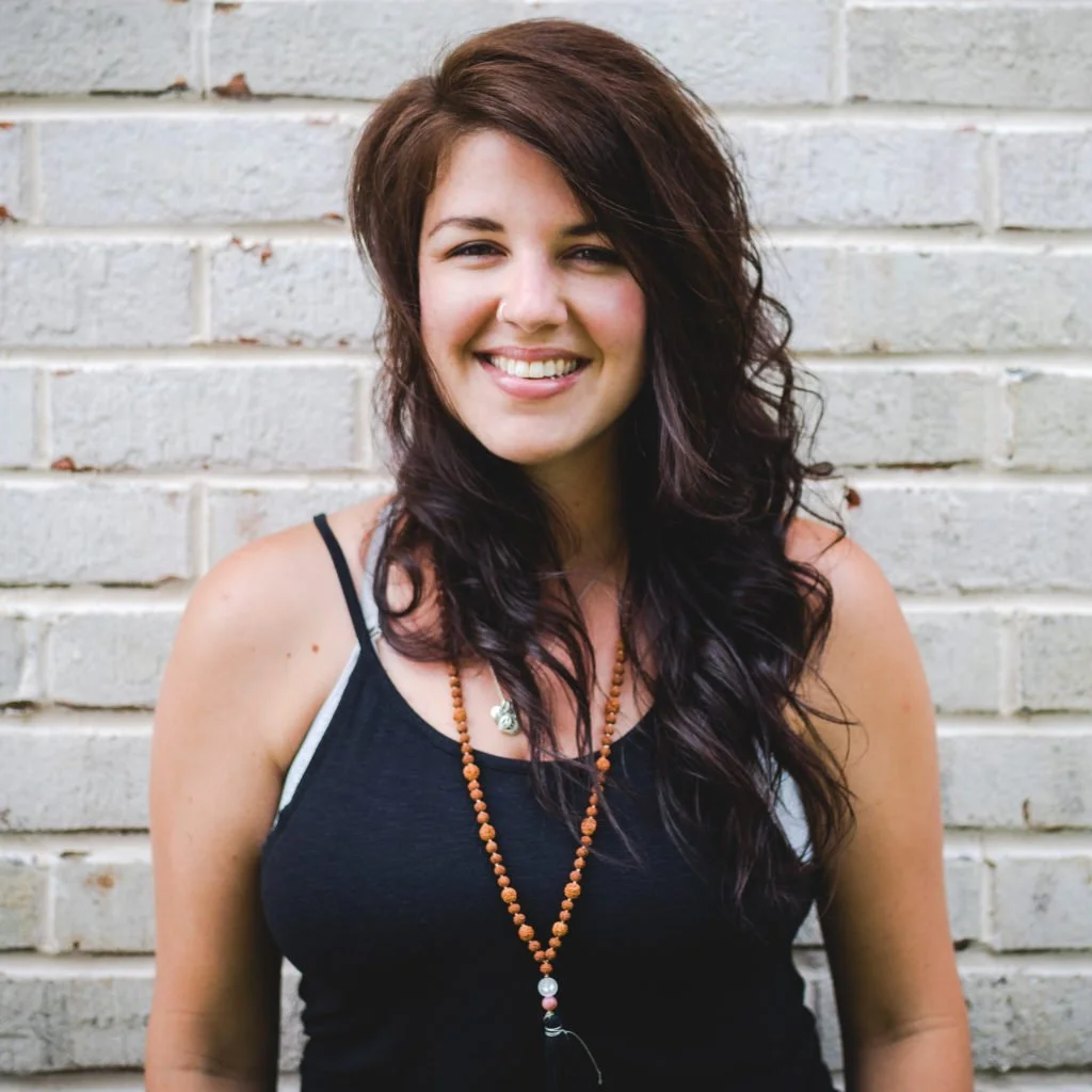A woman smiling in front of a white brick wall, wearing a black tank top and a beaded necklace.