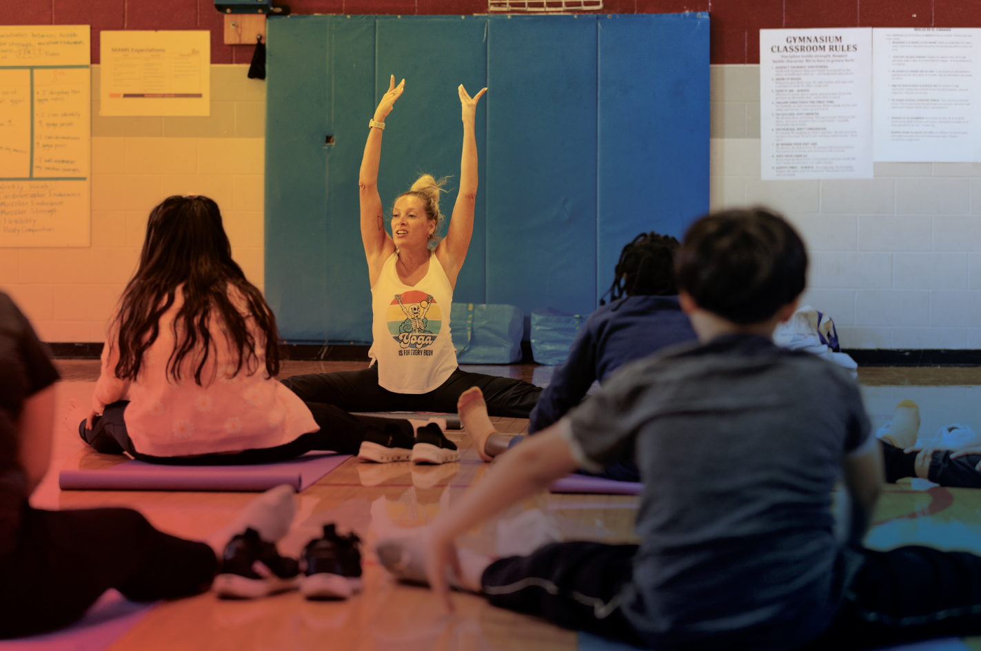 A woman leading a yoga class for children in a gymnasium. She is sitting cross-legged on a mat, with arms raised, demonstrating a pose. The children sit on their mats, attentively watching her. The wall behind features gym rules and colorful mats.
