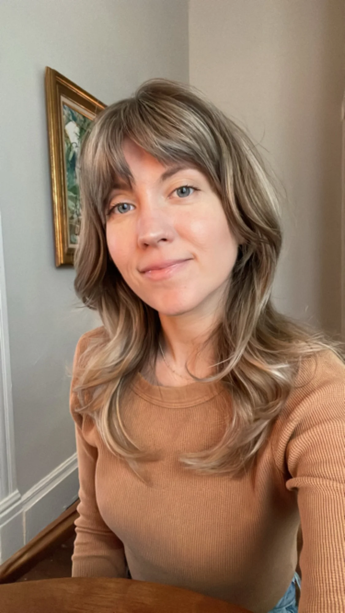 A woman with shoulder-length, wavy, light brown hair with blonde highlights, wearing a tan long-sleeved shirt, sitting at a wooden table indoors. She has light blue eyes and a subtle smile. In the background, there is a gray wall with a colorful framed painting, and a corner of the room with white trim.