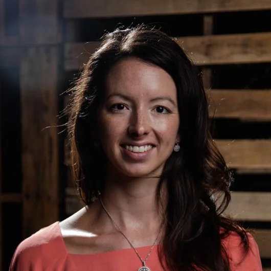 A woman with long dark hair wearing a coral top and necklace, smiling in front of a wooden wall.