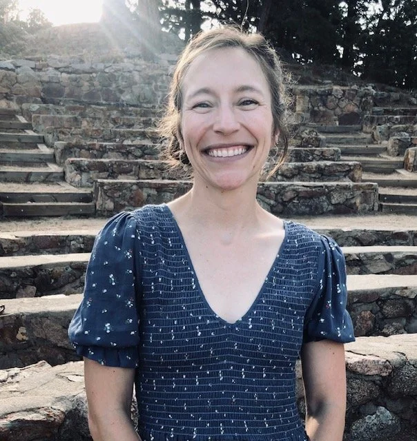 A woman with light skin smiling outside, wearing a blue top with puffed sleeves and a patterned design, standing in front of stone steps and trees.