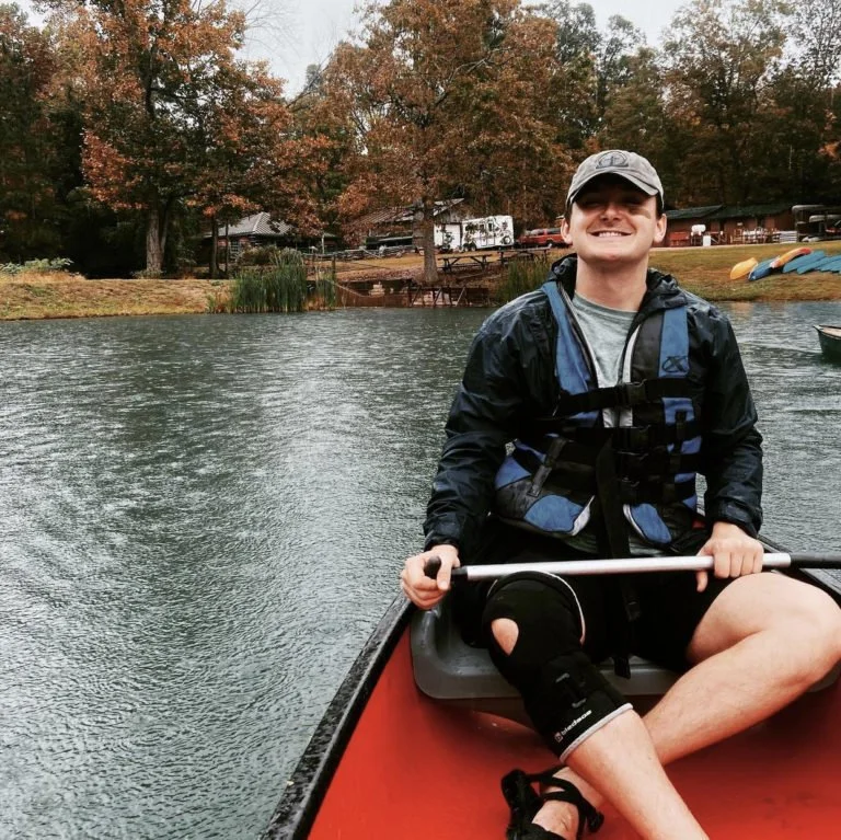 A young man with a knee brace smiling while sitting in a red canoe on a calm lake, surrounded by fall-colored trees.