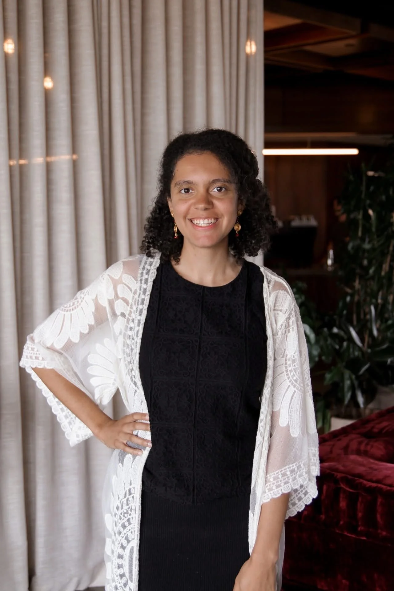 A woman with curly black hair, wearing a black dress and a white lace shawl, standing in a room with beige curtains and dark wood walls, smiling at the camera.