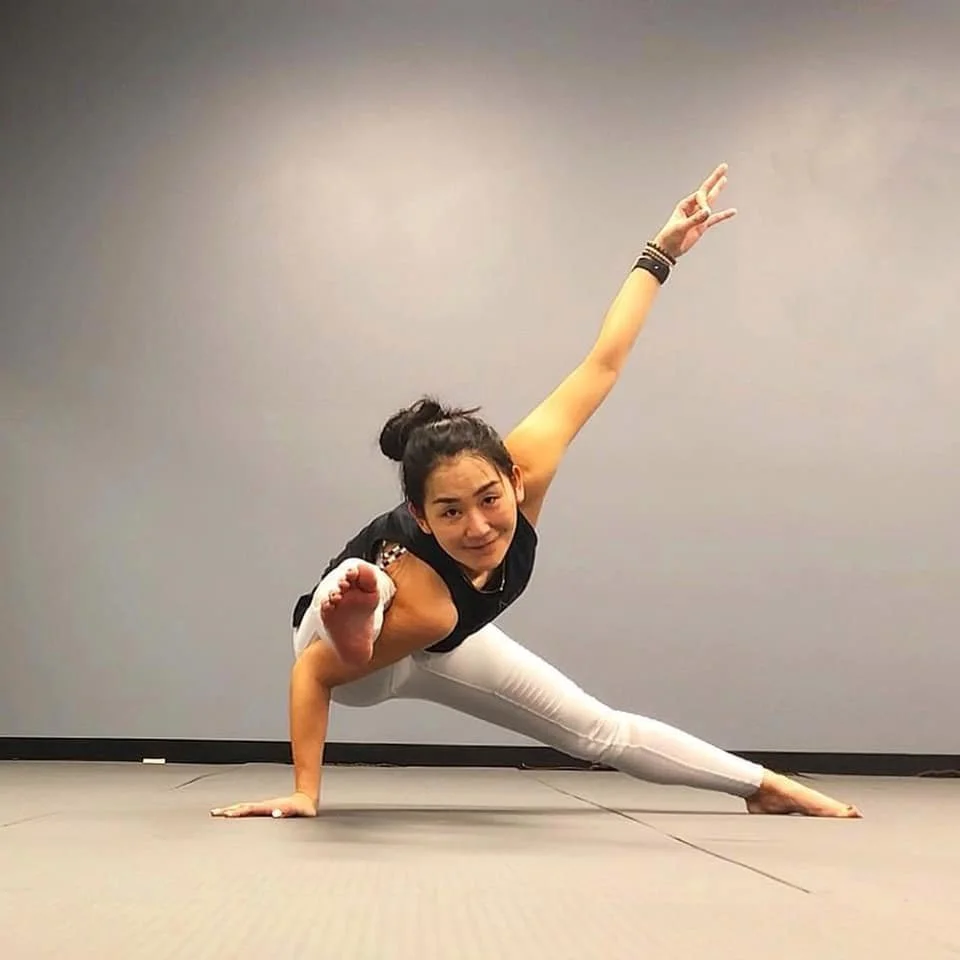Young woman practicing a one-arm side plank yoga pose on a gray floor in a room with plain gray walls.