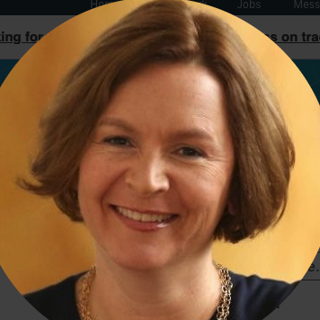 Smiling woman with shoulder-length brown hair, wearing a black top and a necklace, against a yellow background.