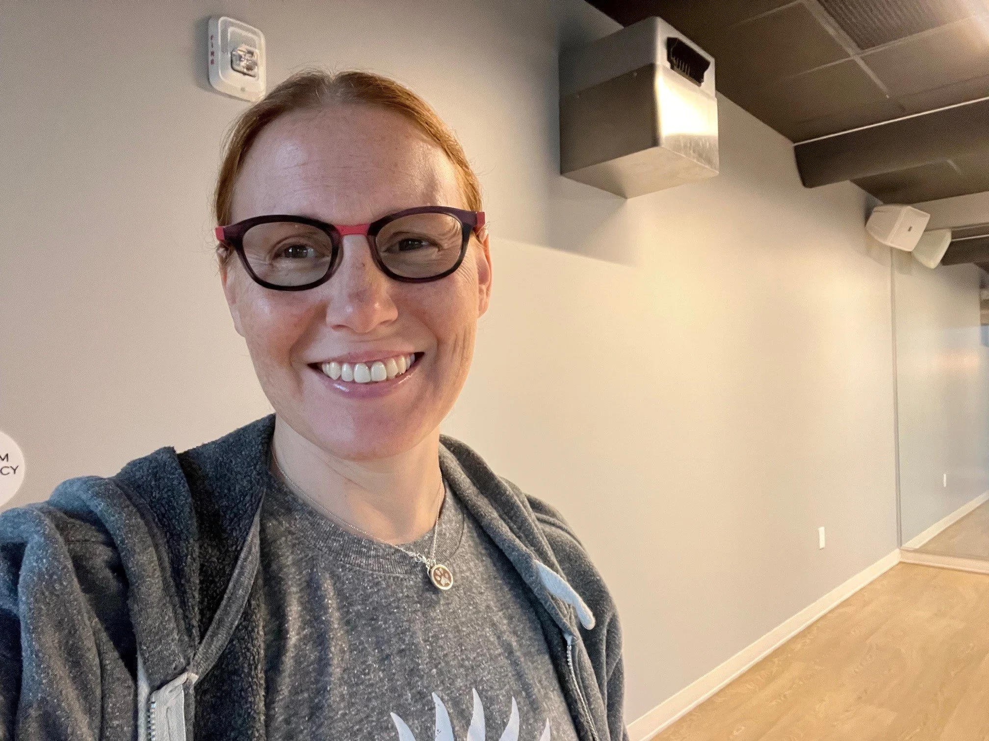 Smiling woman with glasses taking a selfie in an indoor setting with gray walls and modern ceiling fixtures.