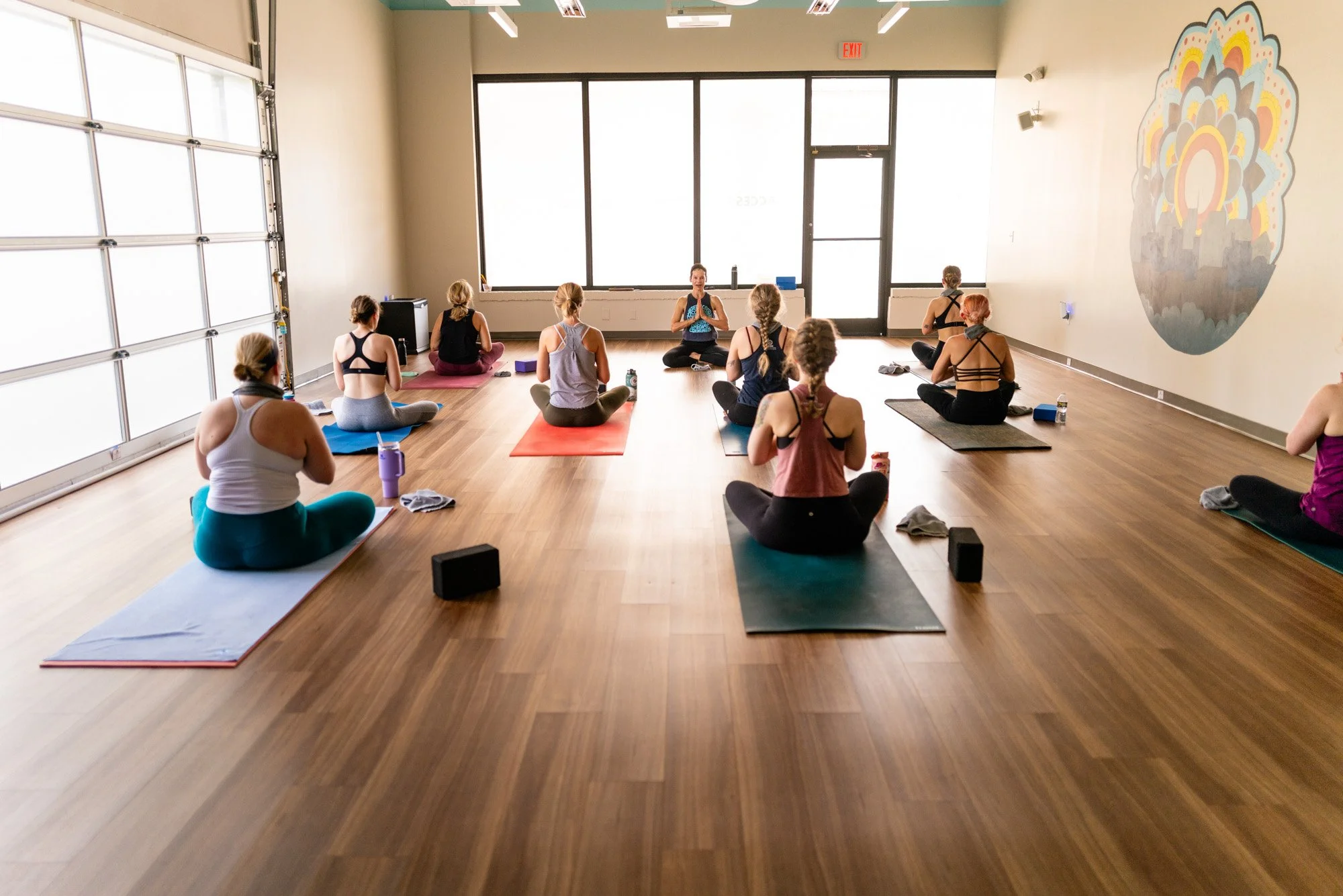 A group of people participating in a yoga class in a spacious, well-lit studio with large windows and wooden floors. The instructor is seated at the front, leading the session, and the students are sitting on yoga mats, listening attentively.