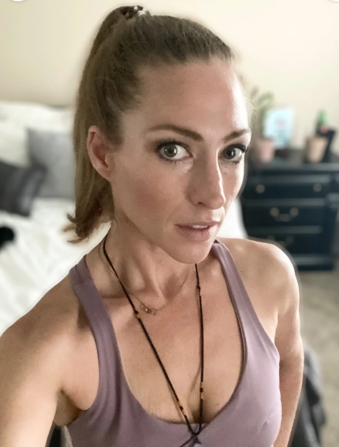 A woman with long brown hair tied back, wearing a purple tank top and layered necklaces, taking a selfie in a bedroom with a bed and dresser in the background.