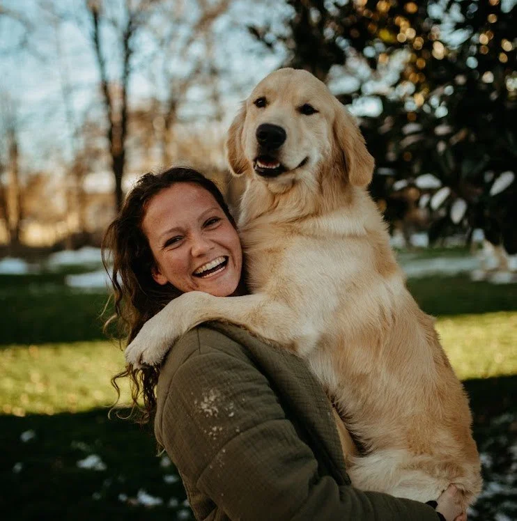 A woman holding a golden retriever puppy outdoors during daytime, smiling and laughing.